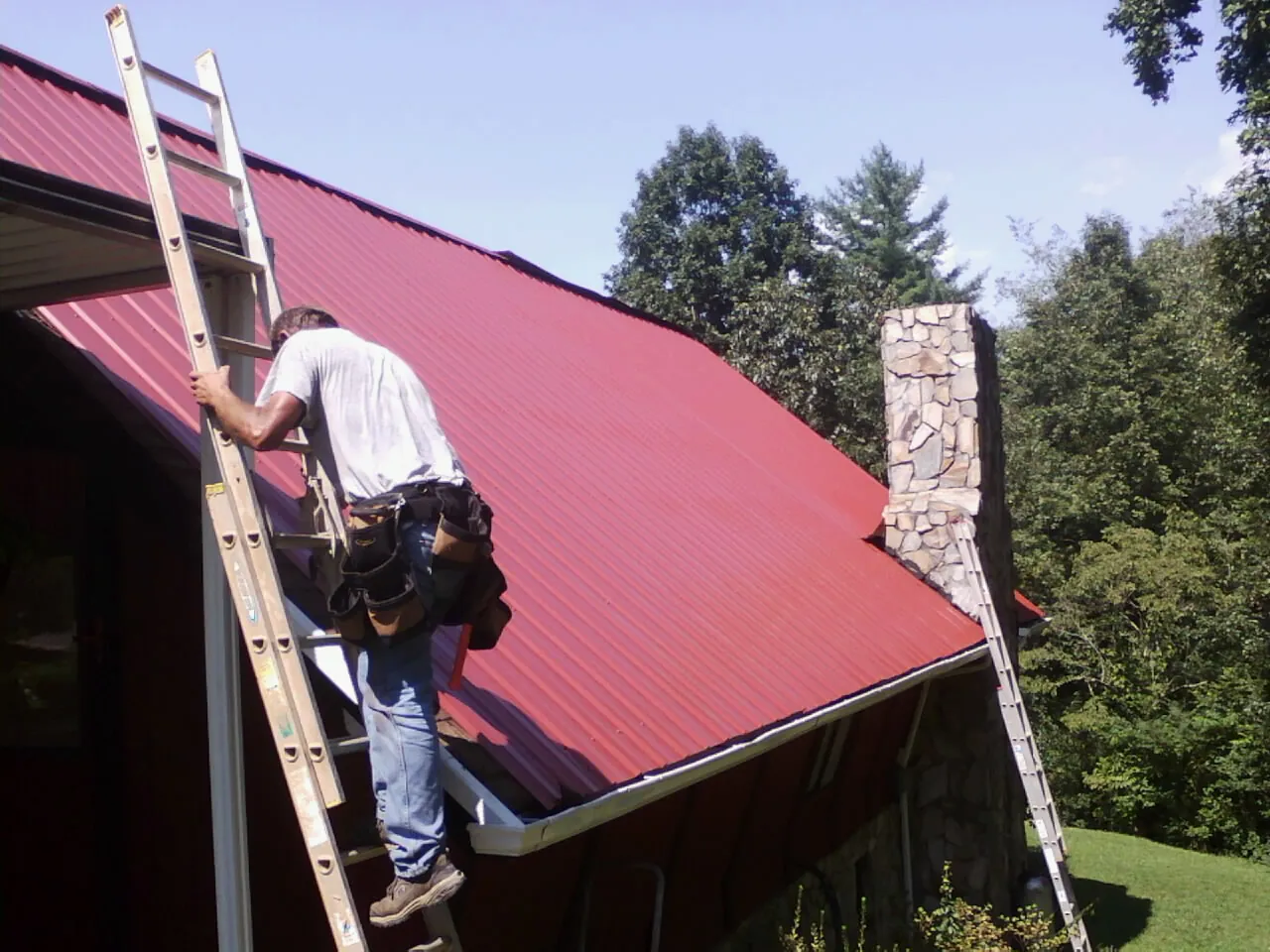 Roofer on a ladder beside a red metal roof and stone chimney — Soffit & Fascia Repair by Roofing Brothers in South Jamesport